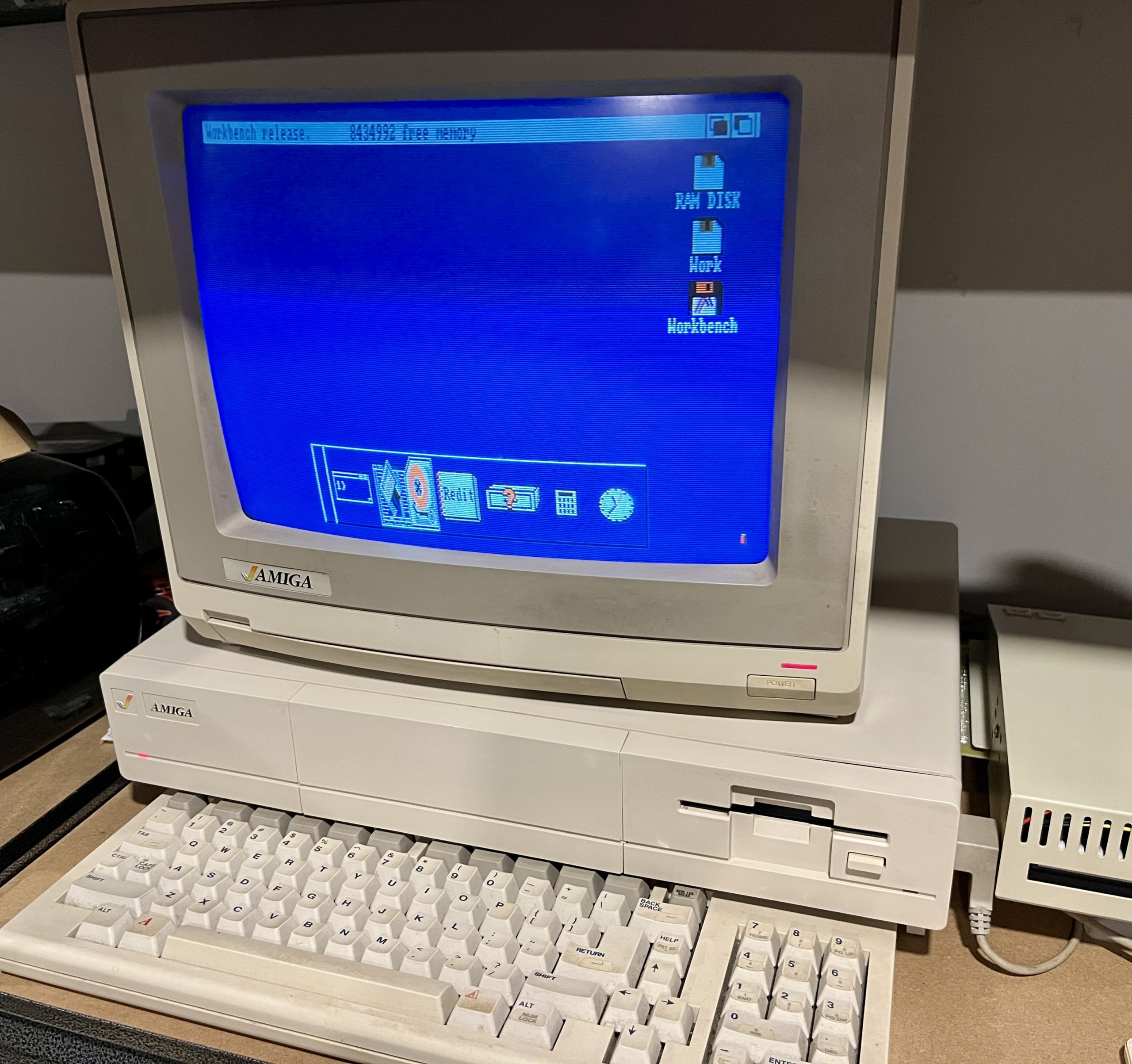 A computer and monitor, a Commodore Amiga 1000 desktop computer with a keyboard in front, an expansion unit attached to the right side, and the Commodore Amiga 1080 monitor on and showing the blue and white Workbench display